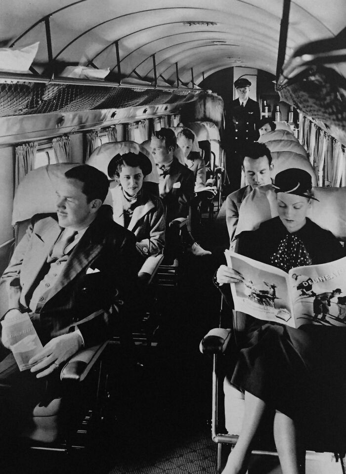 Candid old photos: Passengers seated inside an old airplane cabin, with a flight attendant standing in the aisle.