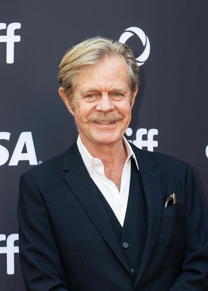 William H. Macy smiling at a film event, wearing a dark suit and white shirt with a mustache and styled hair.