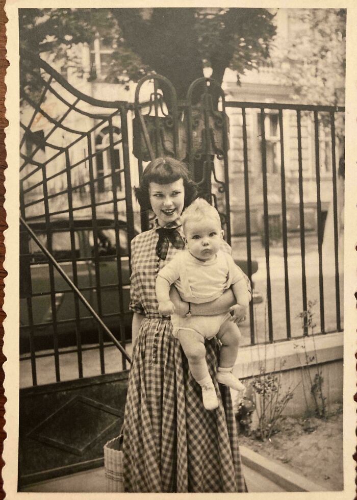 Candid old photos: A young woman in a plaid dress holding a baby, standing in front of an ornate gate.