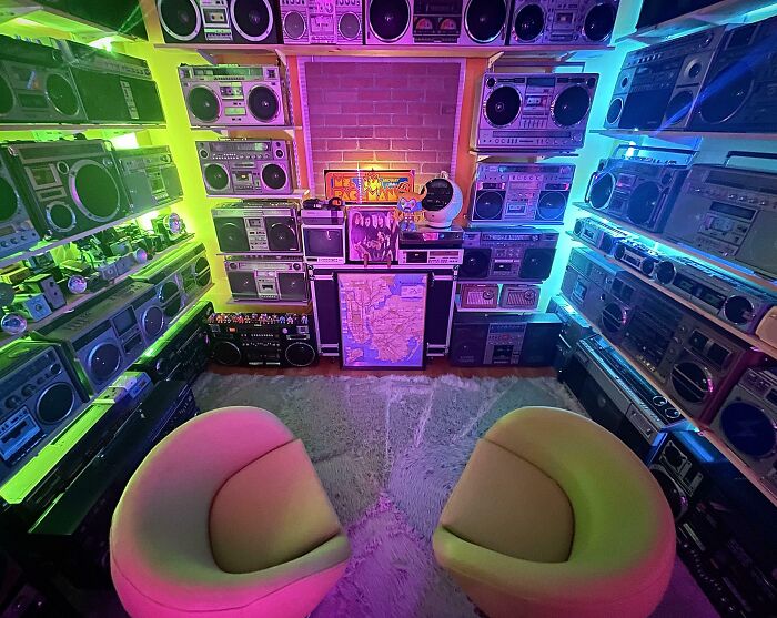 Room filled with a vintage boombox collection, neon lighting, and two modern chairs, showcasing an impressive audio obsession.