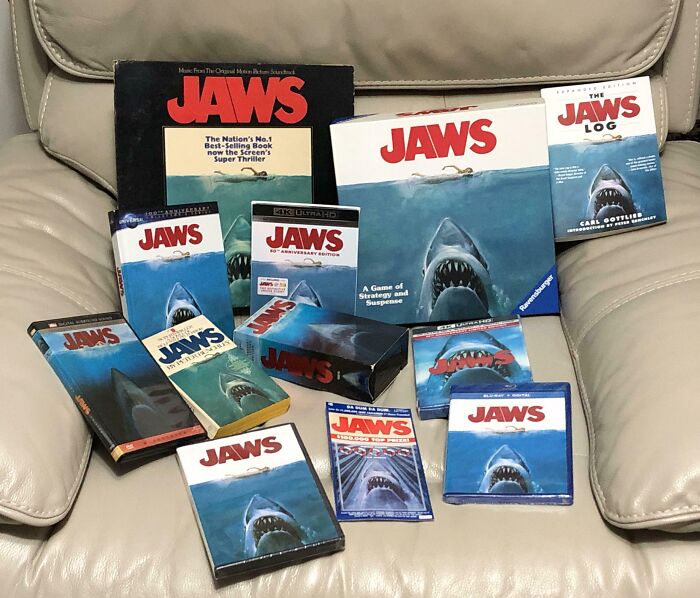 Collection of Jaws memorabilia including DVDs, books, and games displayed on a beige leather couch showing obsession turned collection.