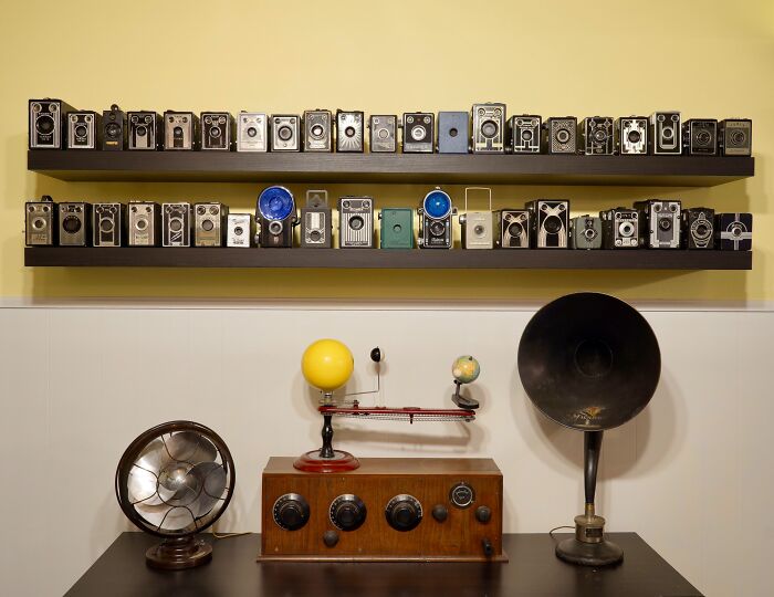 Collection of vintage cameras and antique devices displayed on shelves and a table in a neatly organized room.