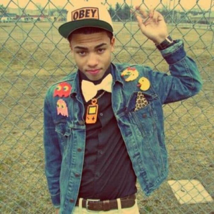 Young man wearing a denim jacket and Obey hat, posing in front of a fence, representing 2010s nostalgia fashion trends.