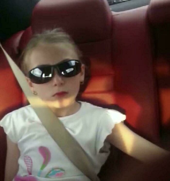 Young girl wearing sunglasses and a white shirt, sitting in a car seat with a seatbelt, 2010s nostalgia vibe.