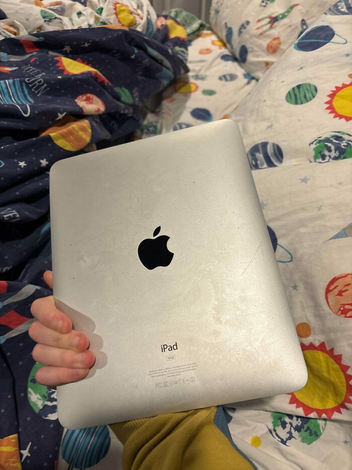Hand holding an older model Apple iPad on a colorful space-themed bedspread, evoking nostalgia for the 2010s.