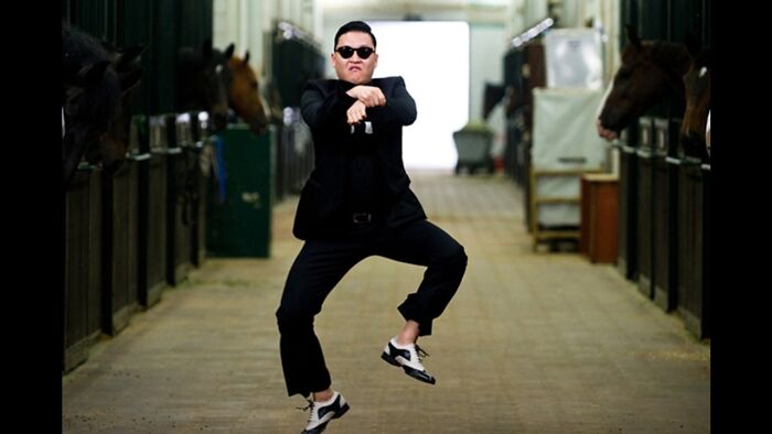 Man in sunglasses and black suit dancing in a stable corridor with horses, capturing 2010s nostalgia vibes.