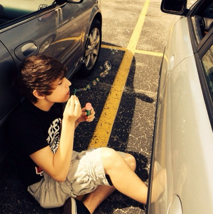 A boy sitting in a parking lot between cars blowing bubbles, capturing a nostalgic moment from the 2010s.