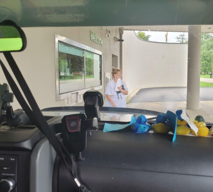 Woman standing outside a building near a welcome sign, viewed from inside a vehicle dashboard, illustrating entitled Karens theme.