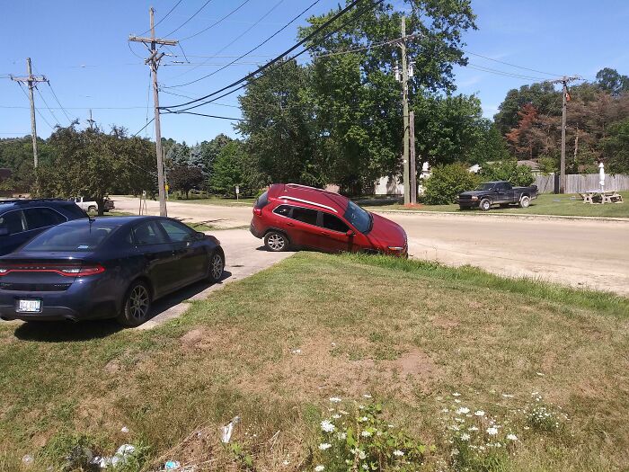 Red car stuck in a ditch on a residential street with other parked vehicles and green trees nearby.