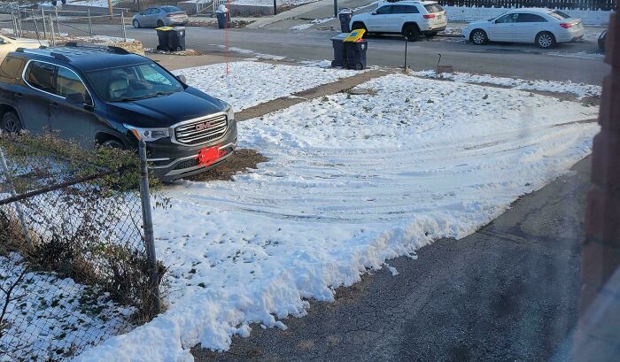 Snow-covered residential driveway with tire tracks, parked cars, and scattered trash bins on a quiet suburban street.