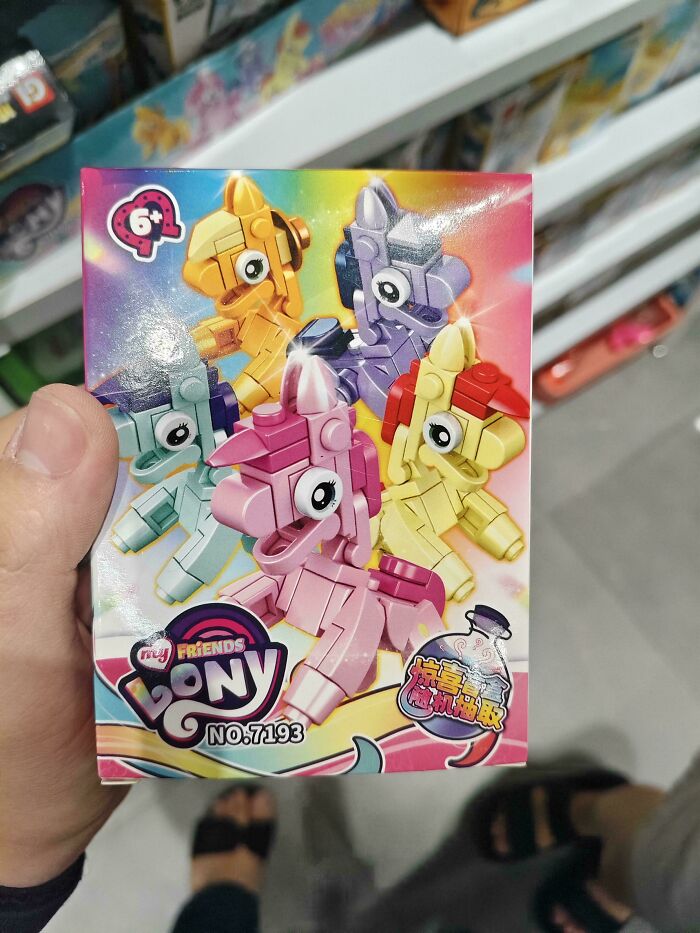 Hand holding a shameless designer toy box featuring colorful copied pony figures from famous brands in a store aisle.
