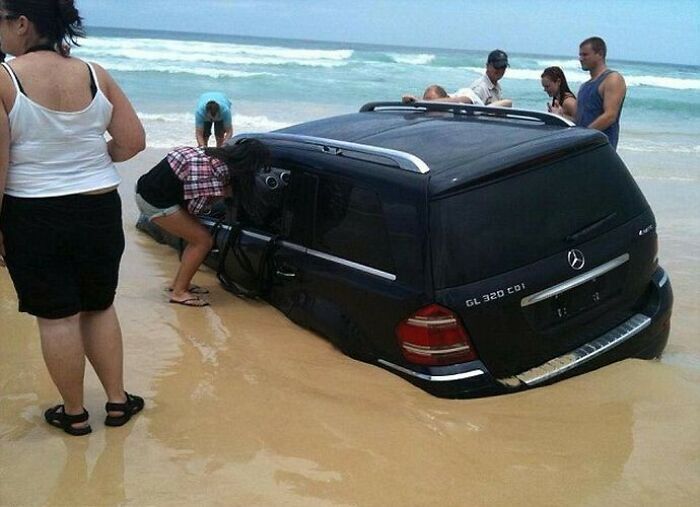 SUV partially submerged in sand at the beach with people inspecting the disaster after forgetting what could go wrong.