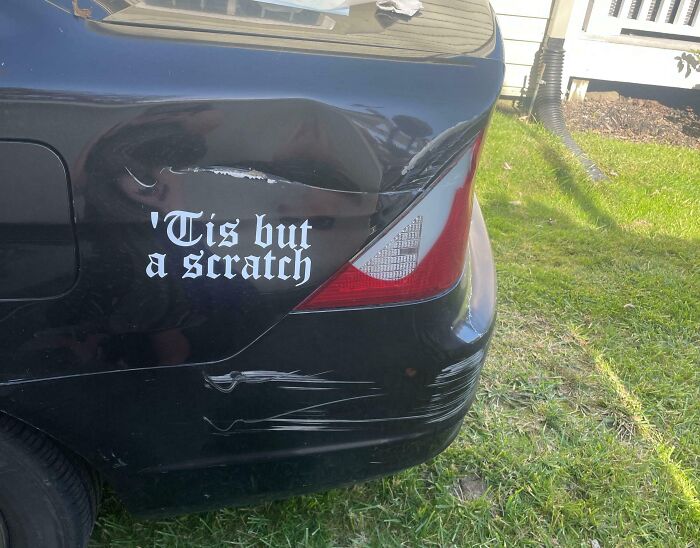 Black car with rear dents and scratches, featuring a humorous decal reading "’Tis but a scratch," showcasing a technically works fix.