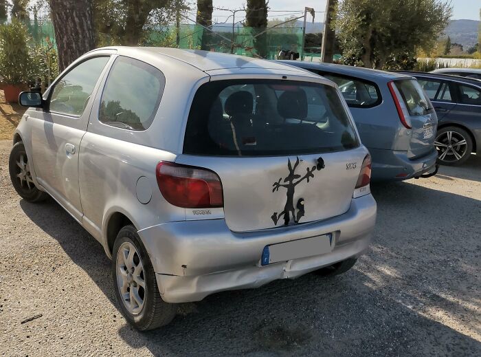 Silver compact car with a damaged rear bumper humorously covered by a painted cartoon figure, showcasing a technically works solution.