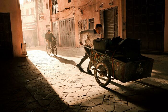 A man pushing a loaded cart down a narrow street in dramatic Accidental Renaissance lighting