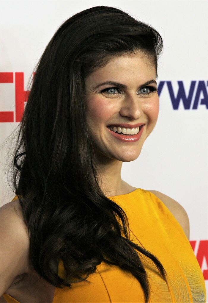 Alexandra Daddario smiling with long dark hair, wearing a yellow dress at a red carpet event.