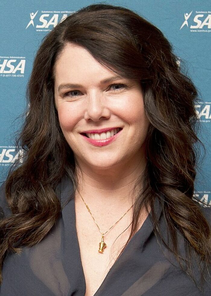 Lauren Graham smiling with long wavy hair, wearing a dark blouse and gold necklace at an event background.