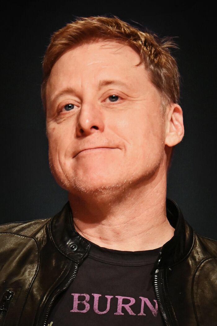 Alan Tudyk close-up portrait wearing a black leather jacket and t-shirt with a neutral expression on a dark background