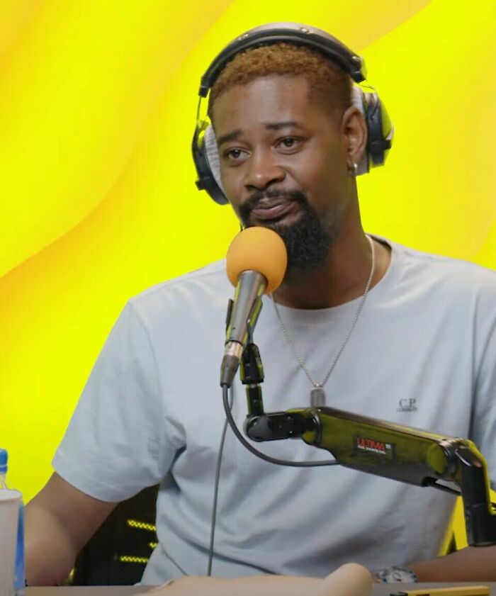 Danny Brown wearing headphones and speaking into a microphone during a podcast recording session with a bright yellow background.