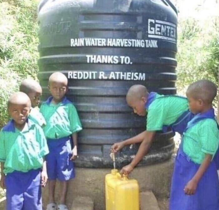 Several kids in green and blue uniforms gather around a rainwater harvesting tank with an unusual donation message.