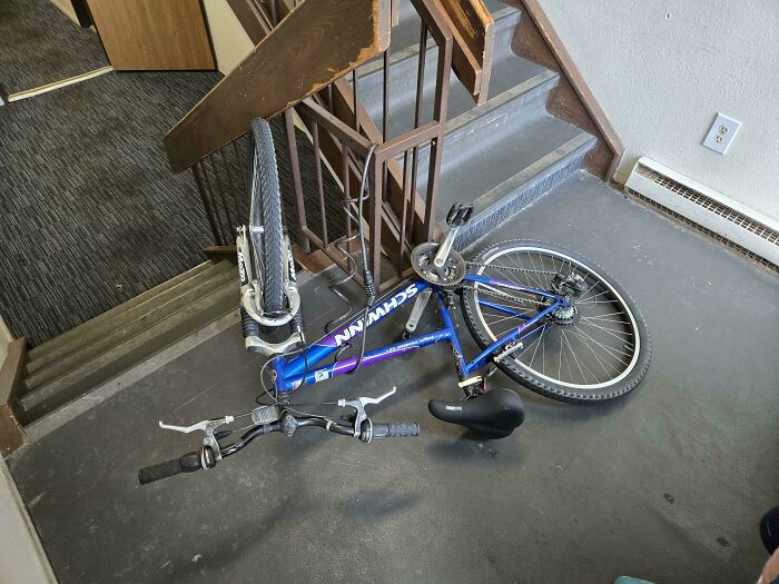 Blue Schwinn bicycle locked awkwardly to stair railing in empty indoor hallway, suggesting unusual neighbor behavior.