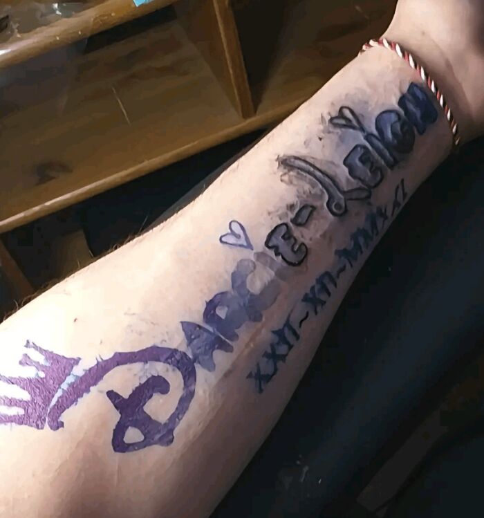 Arm with a truly awful tattoo drawn in marker featuring a crown, hearts, and illegible text in black and purple ink.