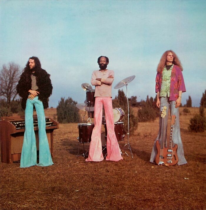 Three musicians in bizarre throwback fashion with wide bell-bottom pants and vintage instruments outdoors.