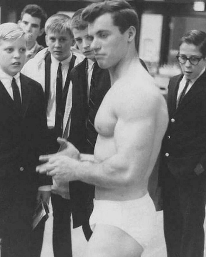 Group of young boys in school uniforms watching a muscular man in vintage underwear in a bizarre throwback moment.