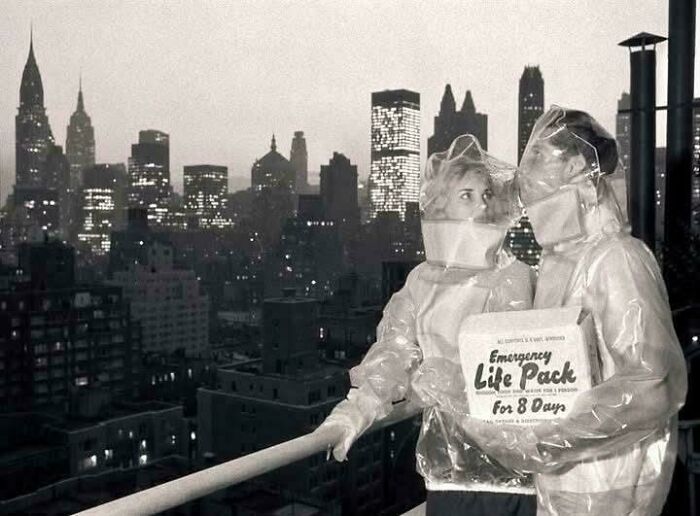 Couple in vintage protective gear holding an emergency life pack against a city skyline in a bizarre throwback moment.