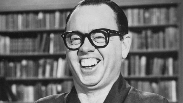 Black and white photo of a man laughing in front of bookshelves showing bizarre throwback moments from the past.