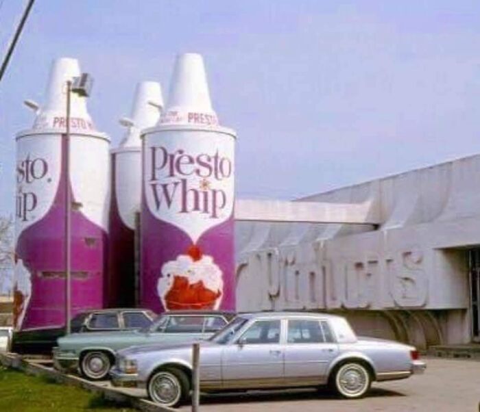 Giant vintage Presto Whip cans outside a building with classic cars in front, showcasing bizarre throwback moments.