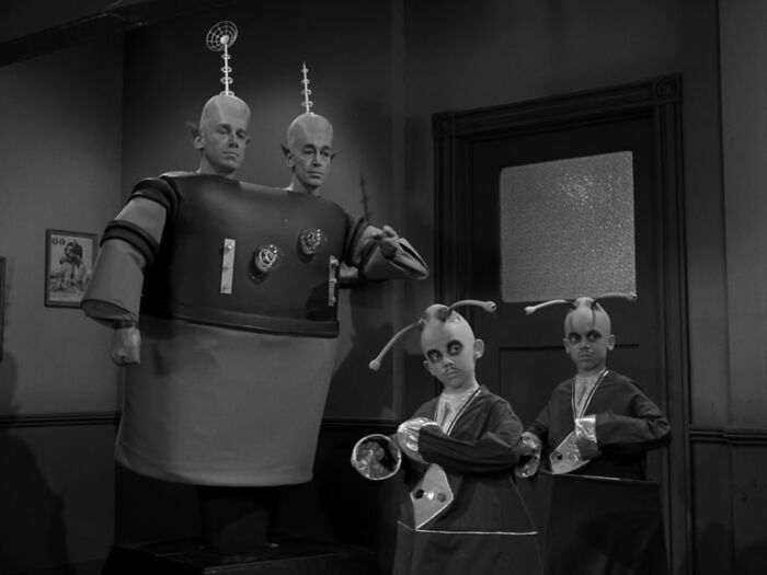 Black and white bizarre throwback moment showing two men and two children in alien costumes with antenna headpieces indoors.