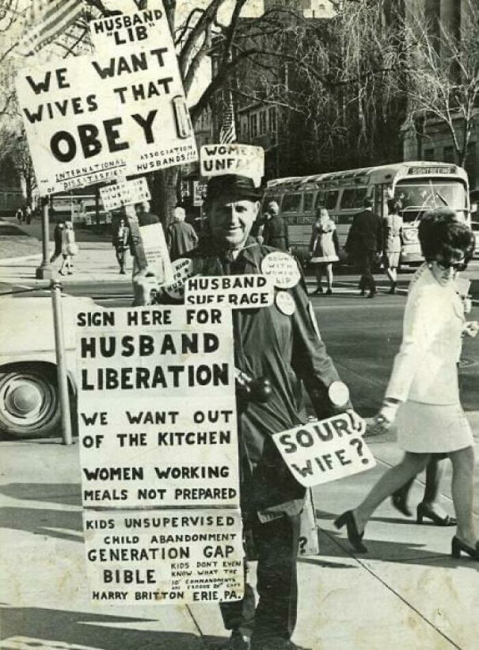 Man holding vintage protest signs about husband liberation and wife obedience in a bizarre throwback moment.