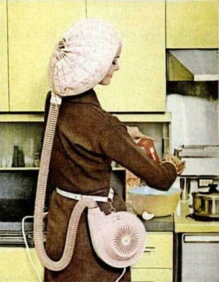 Woman using a bizarre vintage hair drying helmet with attached fan in a retro kitchen, a strange throwback moment.