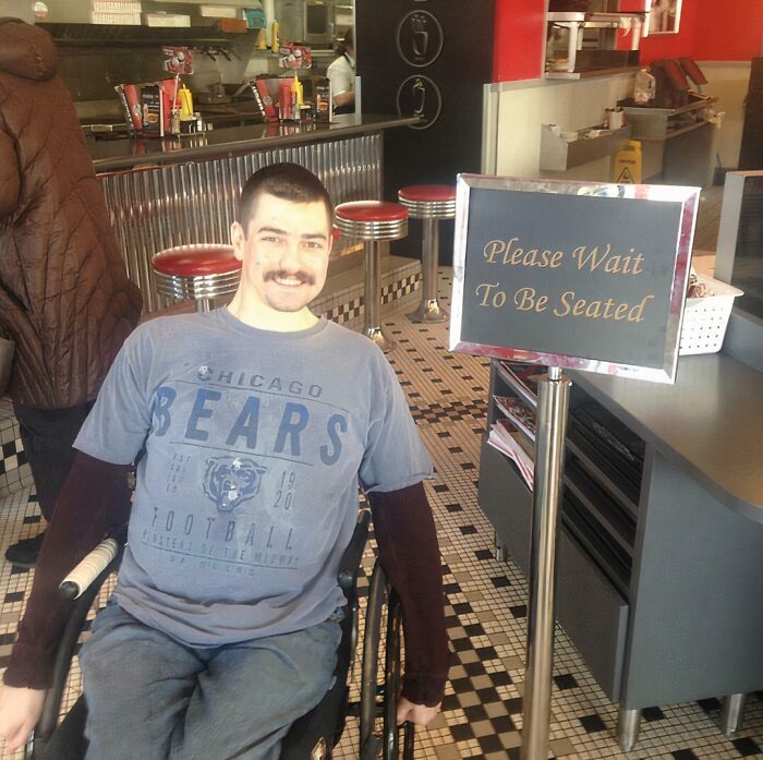 Man in a wheelchair smiling next to a please wait to be seated sign in a diner with sarcastic irony vibe.