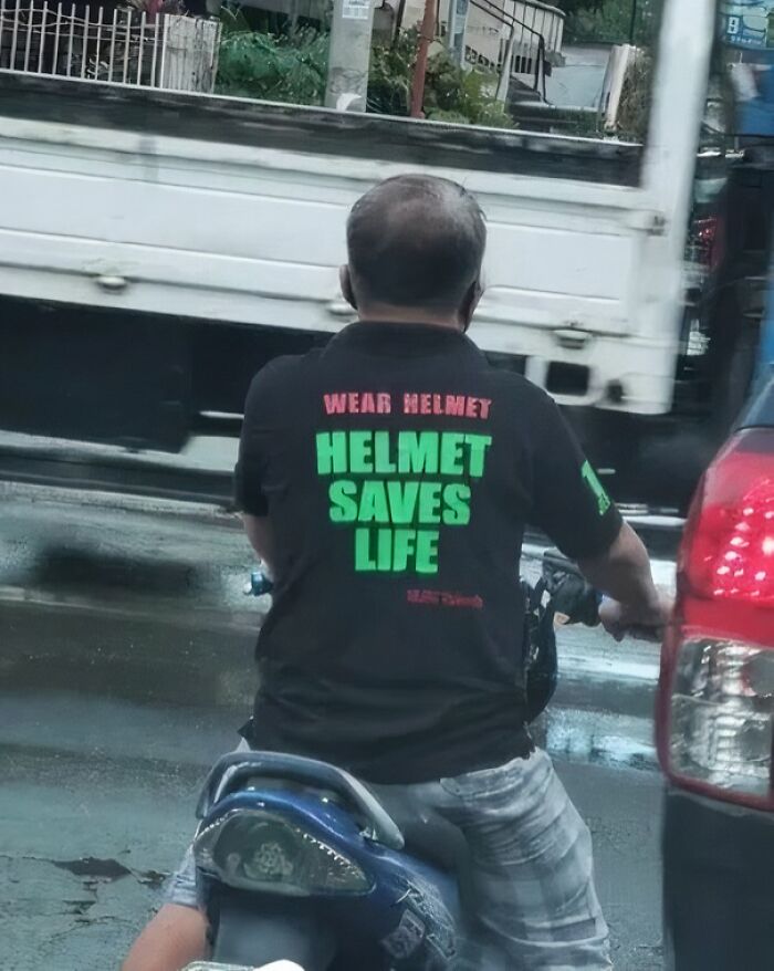 Man riding motorcycle without a helmet wearing a shirt that says helmet saves life in green letters on a busy street.