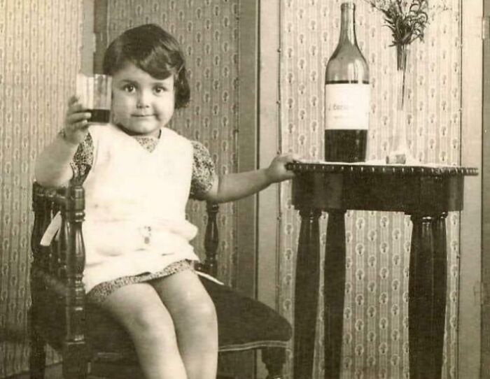 Vintage photo of a young child holding a glass, sitting by a table with a bottle, showcasing bizarre throwback moments.