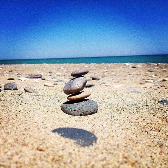 Stacked stones appear to be levitating above sandy beach with ocean and clear blue sky in background, unedited photo.