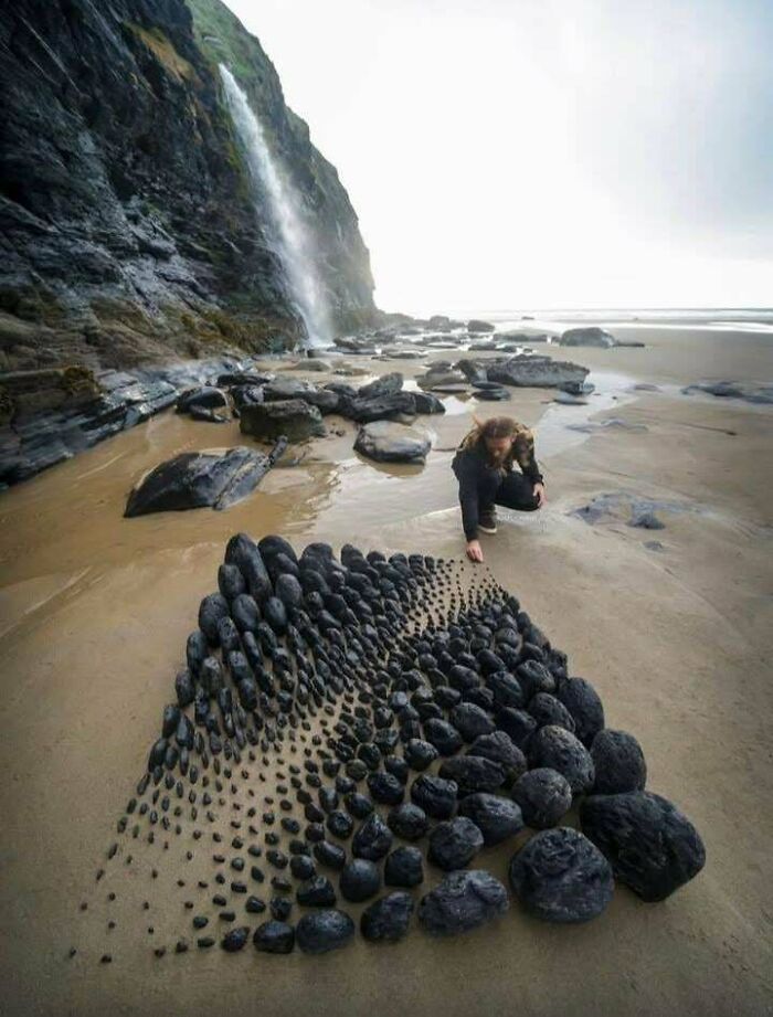 Amazing photos: A person arranging dark stones on a sandy beach next to a waterfall, creating a unique pattern.