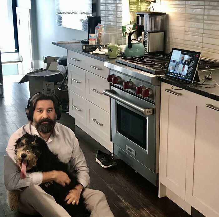 Man sitting on kitchen floor with dog, participating in a virtual meeting, showcasing dads and the pets they love.