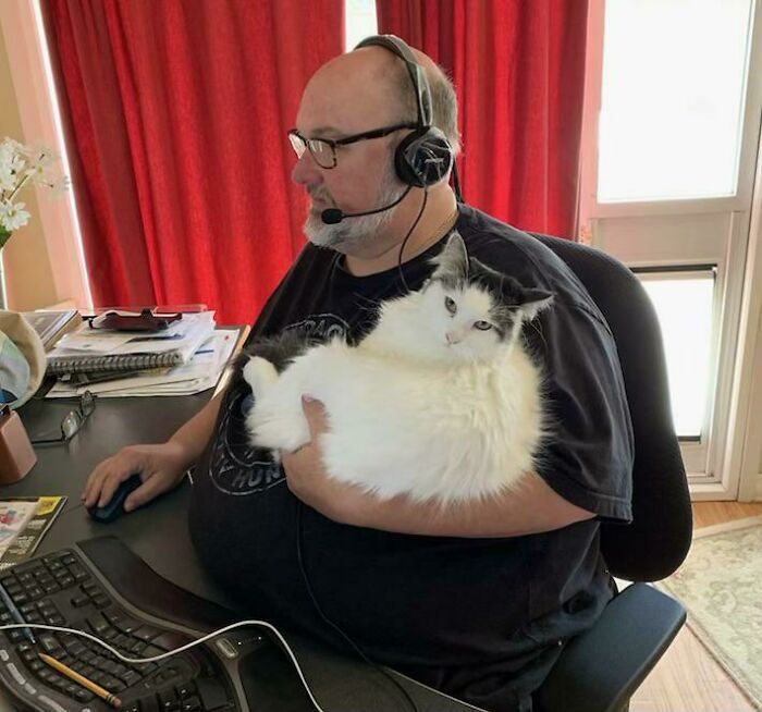 Dad wearing headset holding a fluffy white and black cat while working at a computer, showing love for unexpected pets.