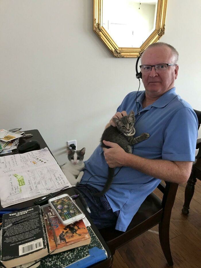 Man wearing headset holding a cat with another cat on his lap showing dads and pets they never wanted but now love