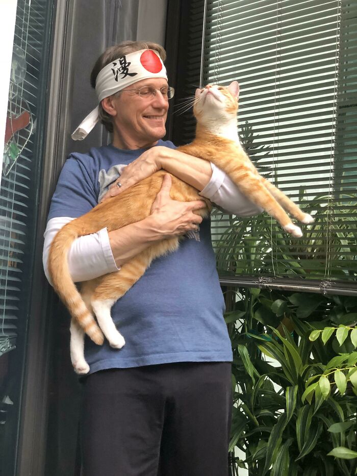 Man wearing a headband happily holding an orange tabby cat, showing the bond between dads and pets they never wanted but love.