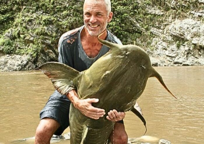 Man holding an enormous strange earth fish with unusual shape in a river surrounded by rocky terrain and greenery.