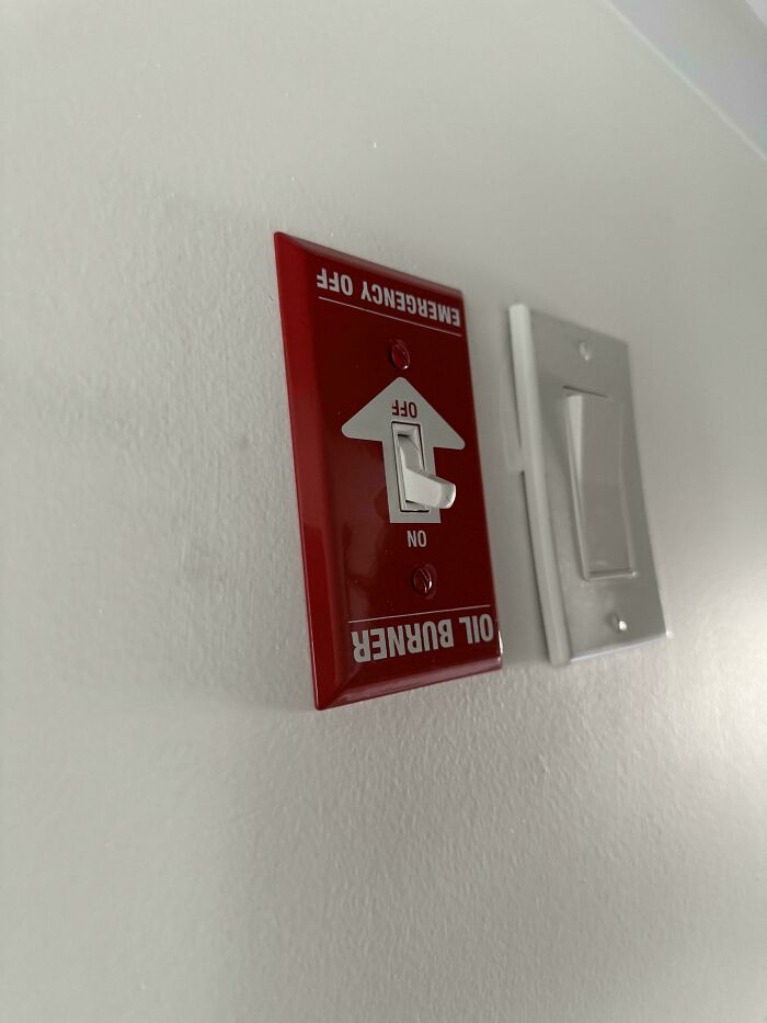 Red emergency oil burner switch next to a white light switch mounted on a plain wall inside a home.