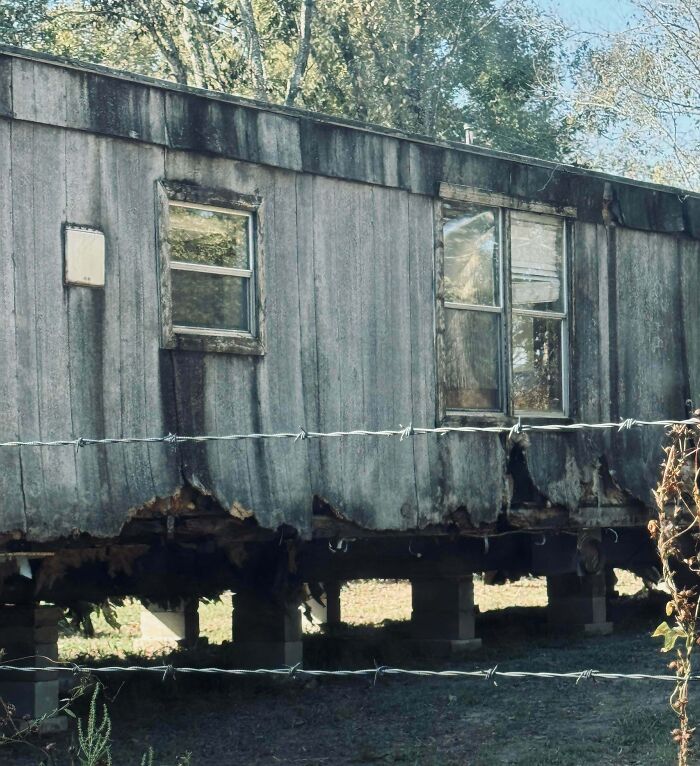 Worn-down trailer home with damaged exterior and broken windows behind barbed wire in a remote area, illustrating neighbors to avoid.