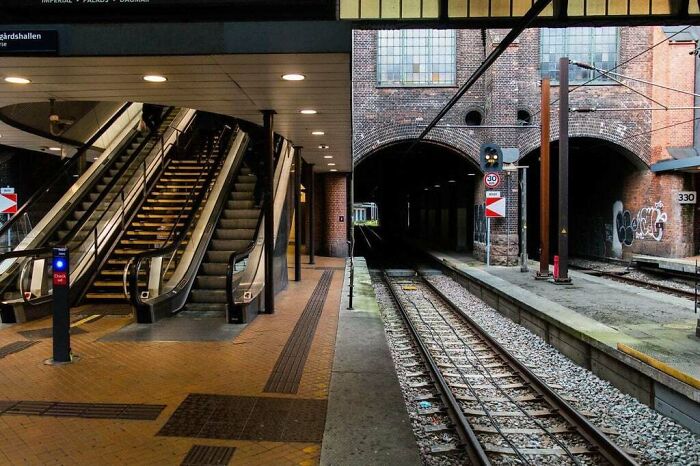 Train station platform with escalators leading up, dark tunnel ahead, and brick walls covered in graffiti.