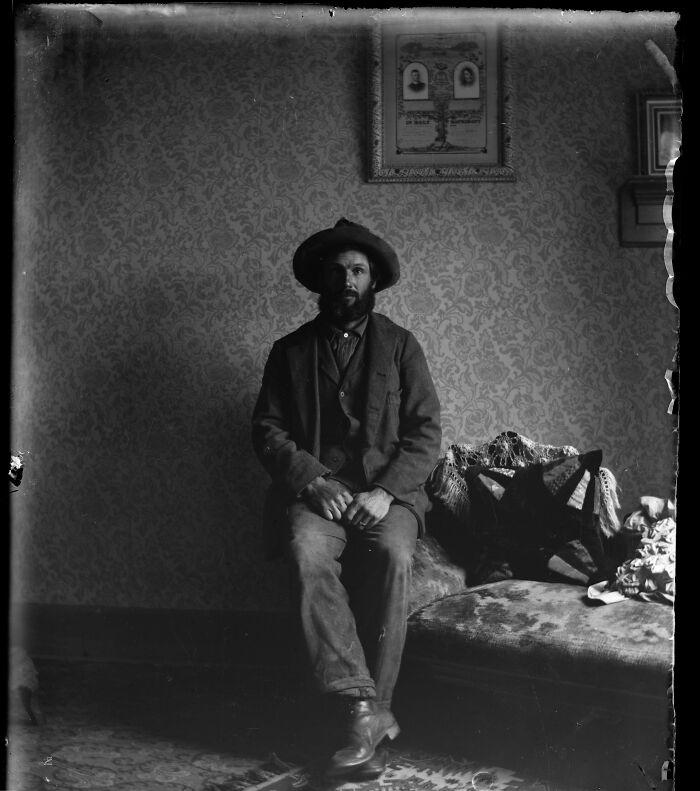 A bearded man in a hat sits on a patterned couch, a candid old photo from generations ago.