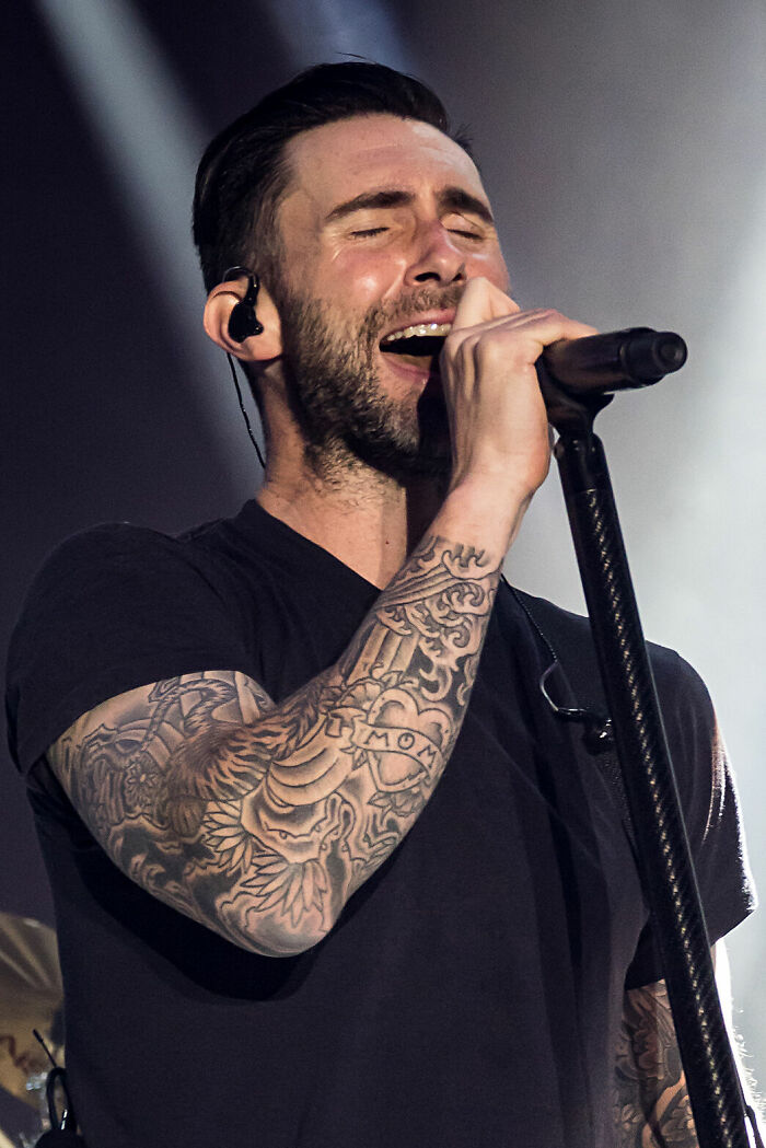 Adam Levine performing live on stage, singing passionately with visible tattoos and wearing a black shirt.