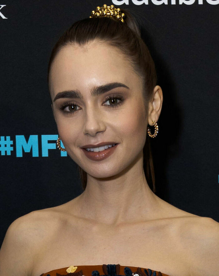 Lily Collins smiling at an event, wearing hoop earrings and a jeweled hair accessory, with natural makeup.